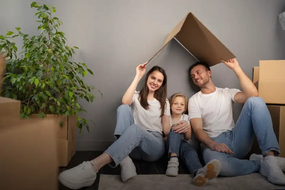 A family sitting among moving boxes, holding a cardboard roof over their heads